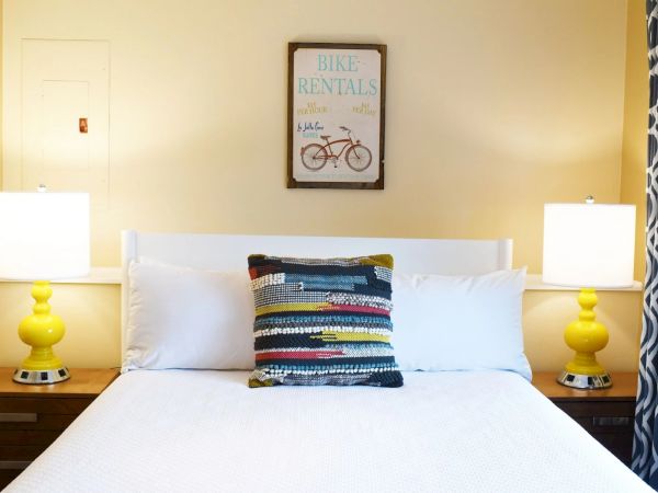A tidy hotel-style bedroom with a white bed, two yellow lamps, a striped throw pillow, and a framed Bike Rentals sign above the headboard, symmetrical lamps.