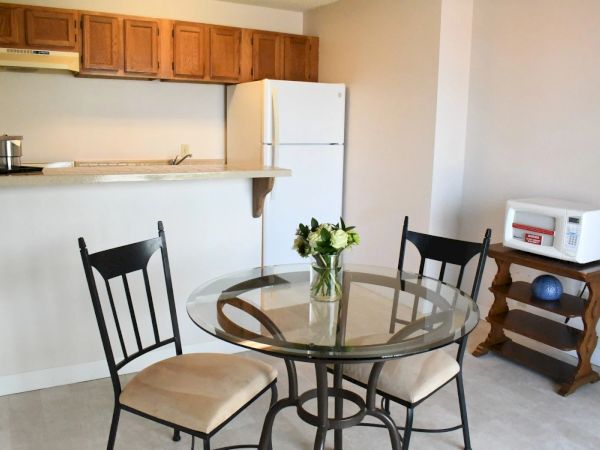 Cozy kitchenette with a glass round dining table, three chairs, a white fridge, wooden cabinets, a small counter bar, and a bouquet of flowers on the table.