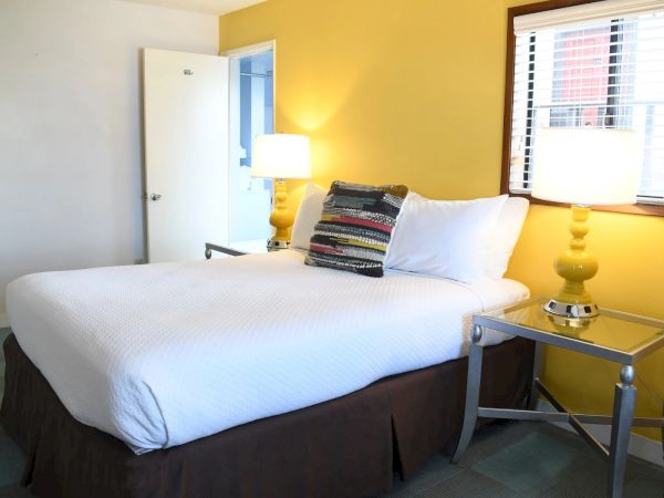A bright hotel room with a white bed, yellow accent wall, two matching bedside lamps, and a glass-topped nightstand on each side.