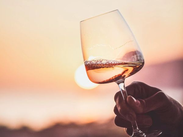 A person holds a wine glass up against a sunset backdrop, the glass catching warm light as the sun dips near the horizon.