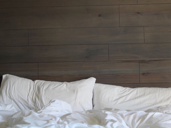 A cluttered bed with white sheets and pillows against a wooden panel wall.