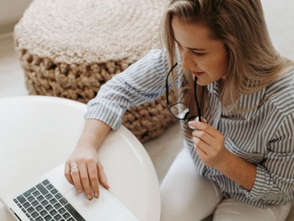 A woman sits at a white table, wearing a striped shirt, looking at a laptop while holding glasses in one hand.