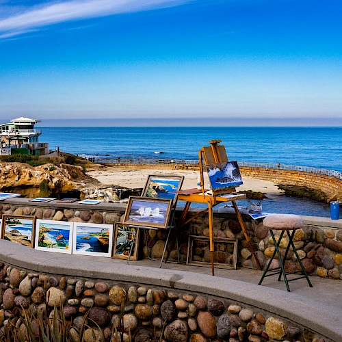 An outdoor seafront gallery with framed photos displayed along a stone wall, a small viewing deck by the sea, and a clear blue sky over calm water.