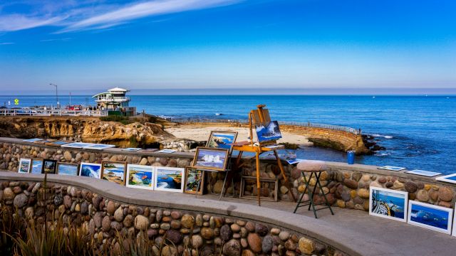 An outdoor seafront gallery with framed photos displayed along a stone wall, a small viewing deck by the sea, and a clear blue sky over calm water.