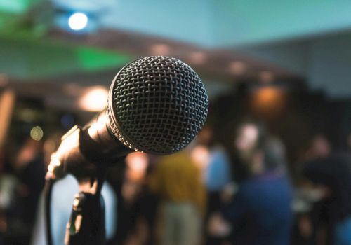 A close-up microphone on a stand in focus, with a blurred crowd and stage lights in the background.