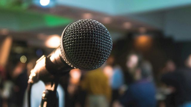 A close-up microphone on a stand in focus, with a blurred crowd and stage lights in the background.