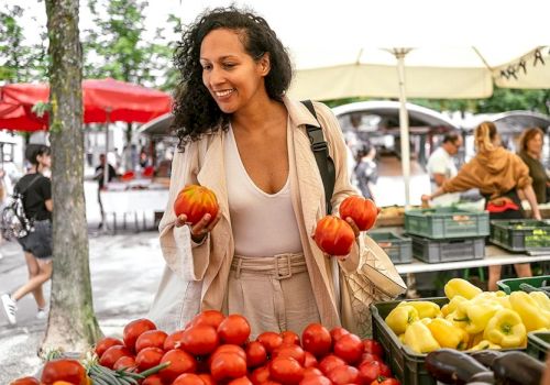A woman shops at a vibrant outdoor market, picking red tomatoes and holding two small pumpkins, with lemons and more produce on display.