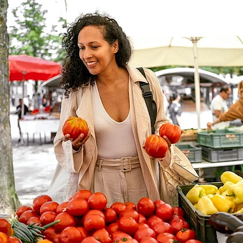 A woman shops at a vibrant outdoor market, picking red tomatoes and holding two small pumpkins, with lemons and more produce on display.