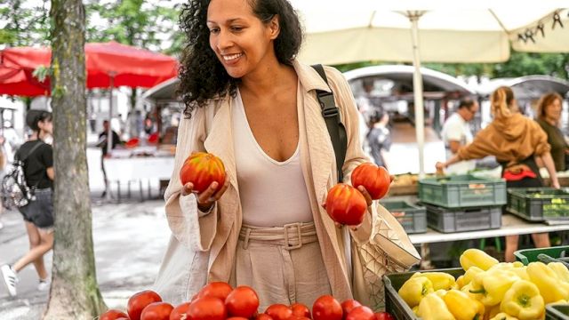 A woman shops at a vibrant outdoor market, picking red tomatoes and holding two small pumpkins, with lemons and more produce on display.