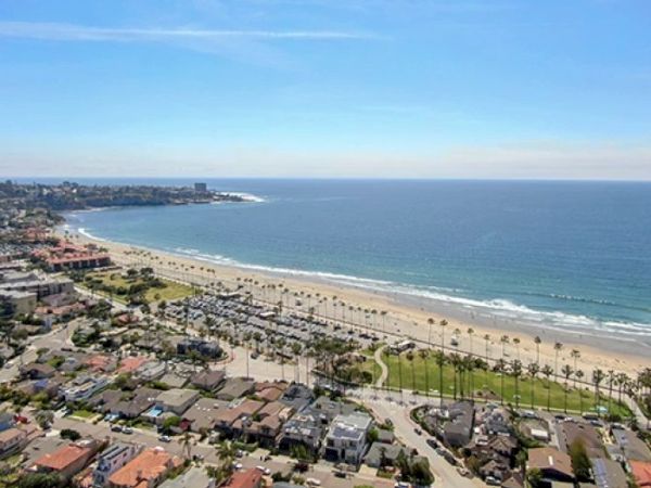 Aerial view of a sunny coastal city: beach, palm-lined promenade, and dense residential blocks along the shoreline, with blue ocean extending far.
