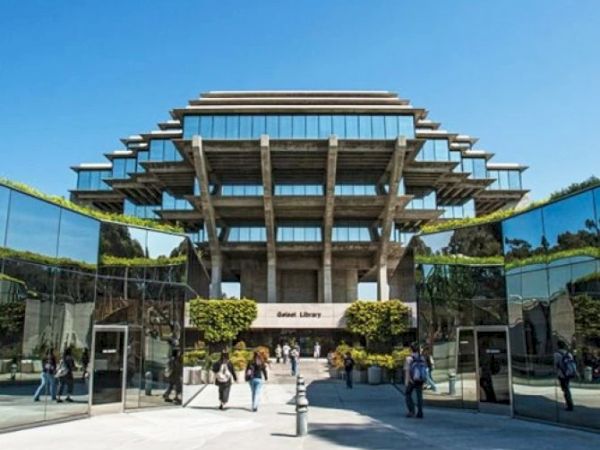 A striking modern glass-and-concrete building with layered, stepped architecture; people walk through a glass-framed entrance on a sunny day.