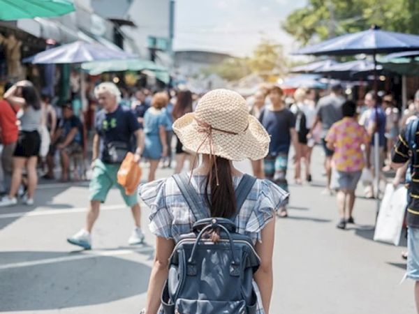 A person with a backpack and straw hat stands in a crowded street market, shoppers and stalls around on a sunny day.