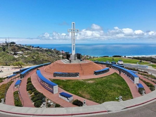 A circular memorial with a large cross at its center atop a terraced platform, overlooking the sea, blue skies, and manicured paths.