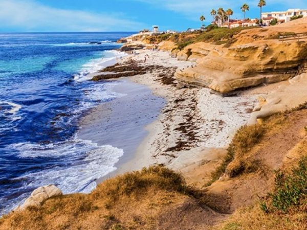 A rugged coastal scene with a sandy cliff edge, blue ocean waves, and dry grassy foreground under a clear sky. Top it at 140 characters, always ending the sentence.