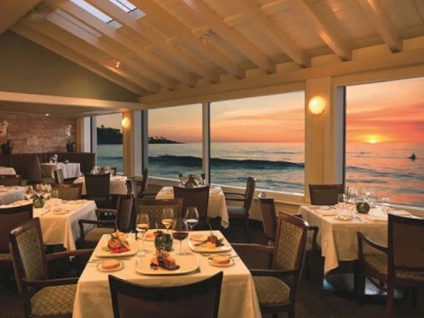 A sunset dining room with ocean view, elegant tables set with white linens, dinnerware, and candles, soft lighting, and balconies over the water.