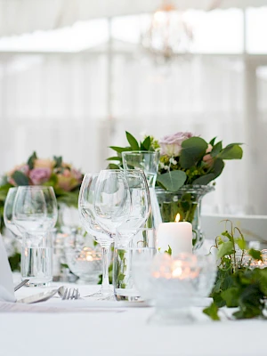 A formal dining table setup with white tablecloths, glassware, candles, and greenery centerpiece in a bright, elegant venue.