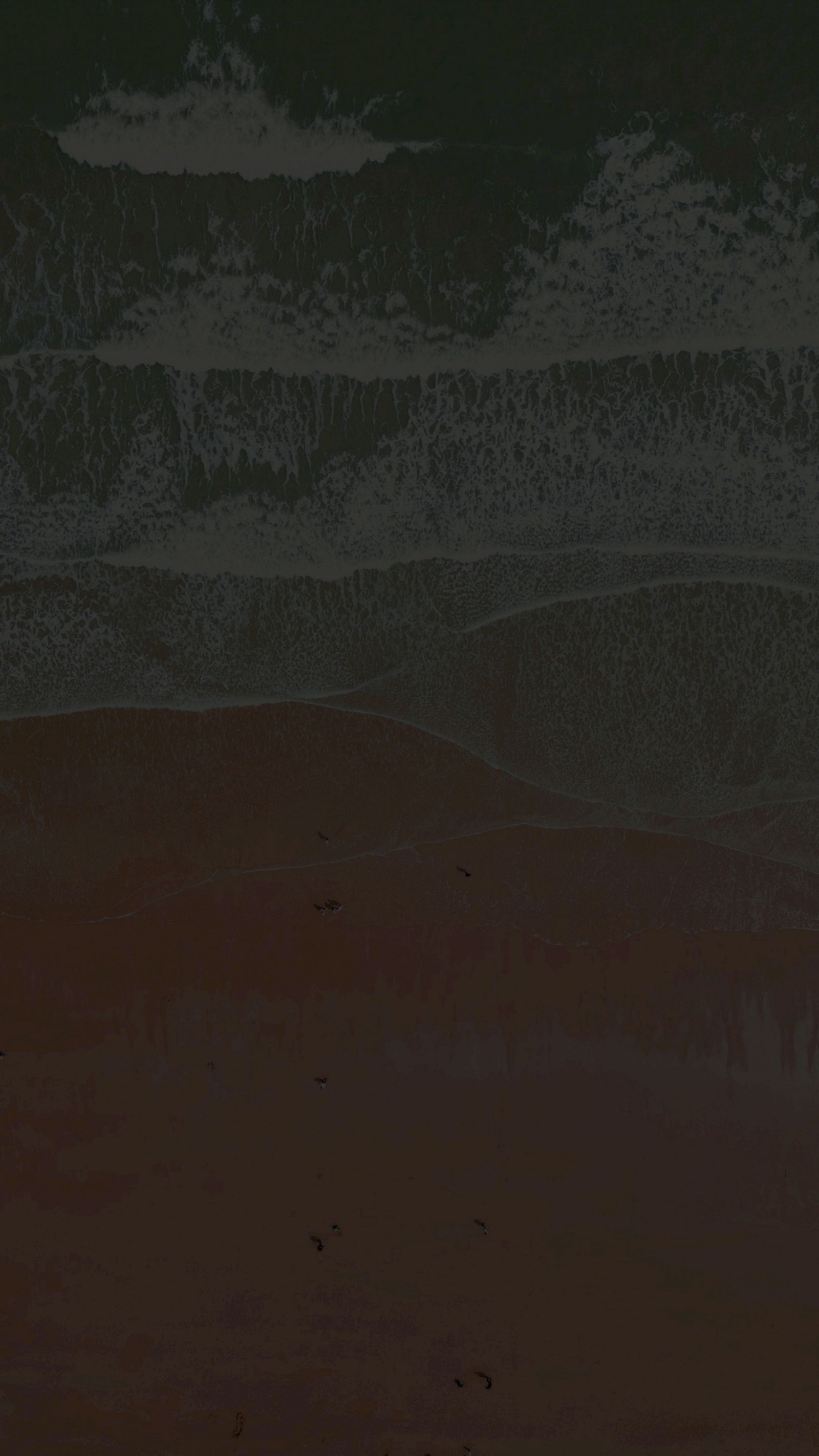 A dark beach scene with foamy waves washing onto the sand, the water&rsquo;s edge fading into shadow at the top, ending in a dim horizon.