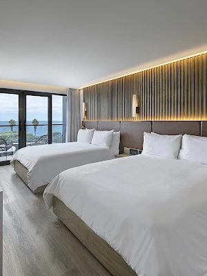 A modern hotel room with two large beds, a sleek desk, ambient wall lighting, and a balcony with a scenic view beyond.