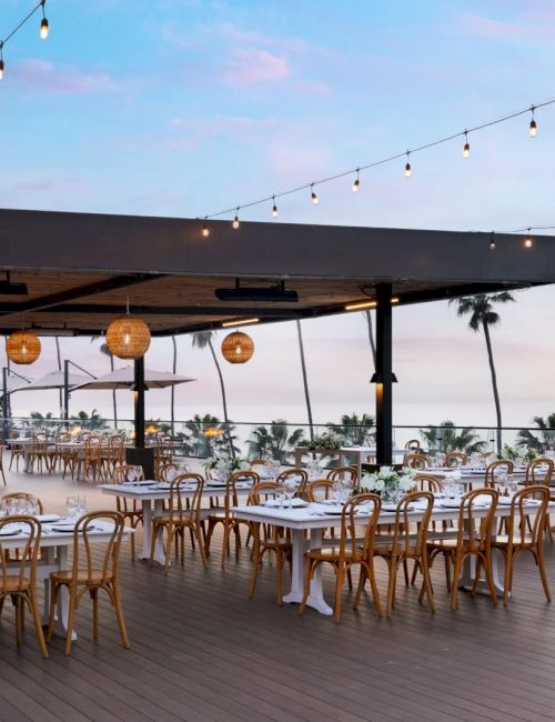 A stylish outdoor dining area with many wooden chairs and white-table settings under string lights, facing a sunset over palm trees and the ocean.