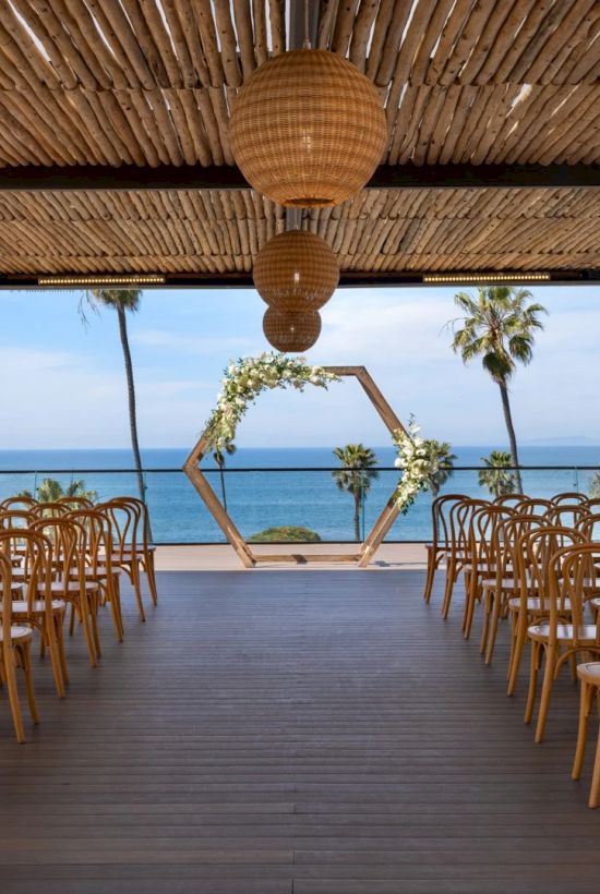 A beach wedding setup with wooden chairs, a driftwood aisle, a hexagonal arch, and palm trees by the ocean under a thatched roof canopy, all facing the sea.