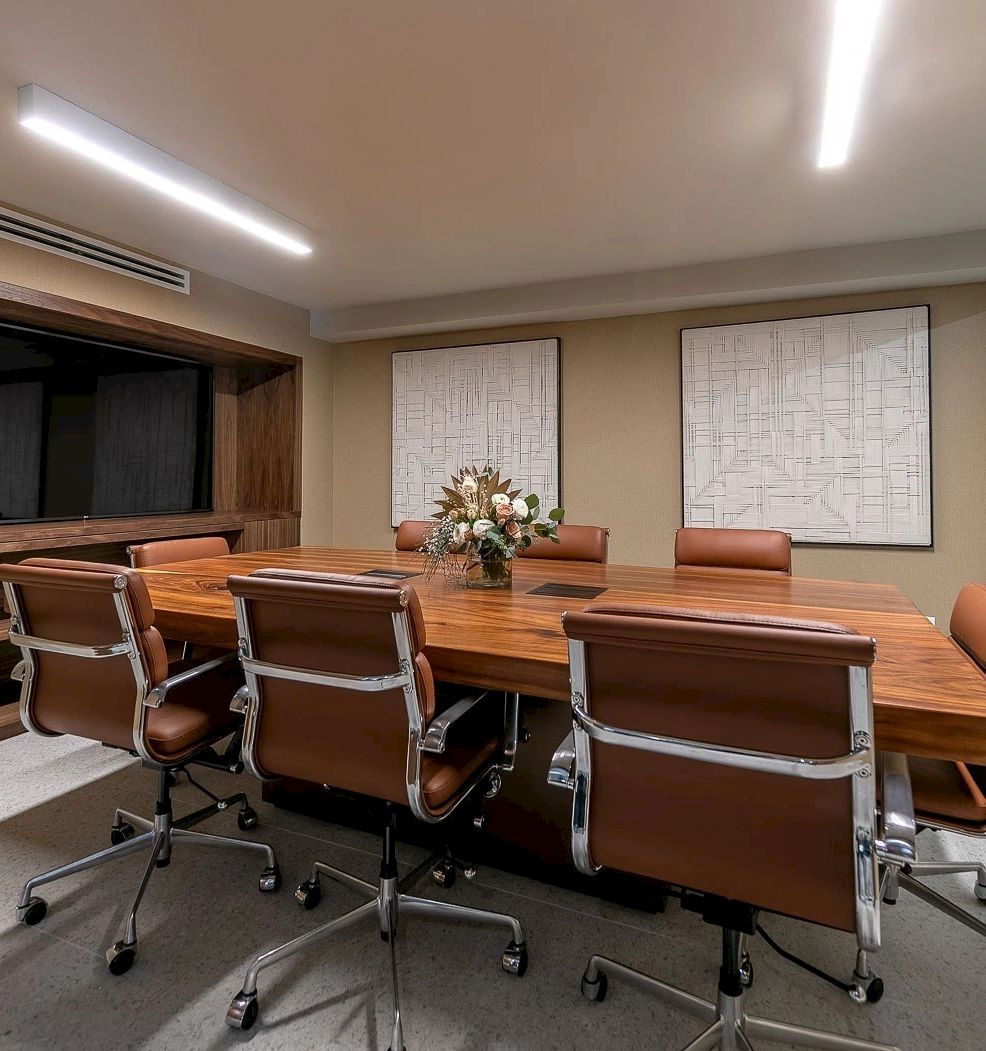 A modern conference room with a long wooden table, brown leather chairs, a large flat screen, decorative flower arrangement, and glass wall to an adjacent area.