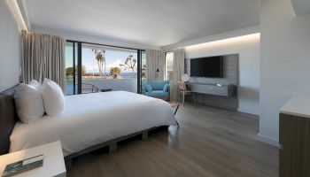 A modern hotel room with a large bed, light wood floors, a wall-mounted TV, a seating area, and a balcony view of palm trees and the water, bright and airy.