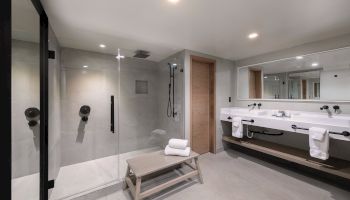 A modern, spacious bathroom with a glass walk-in shower, double vanity, large mirror, and a small bench in front of the shower.