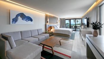 A modern hotel suite with a gray sectional sofa, wooden coffee table, king bed, artwork, and a balcony view with plants and natural light.