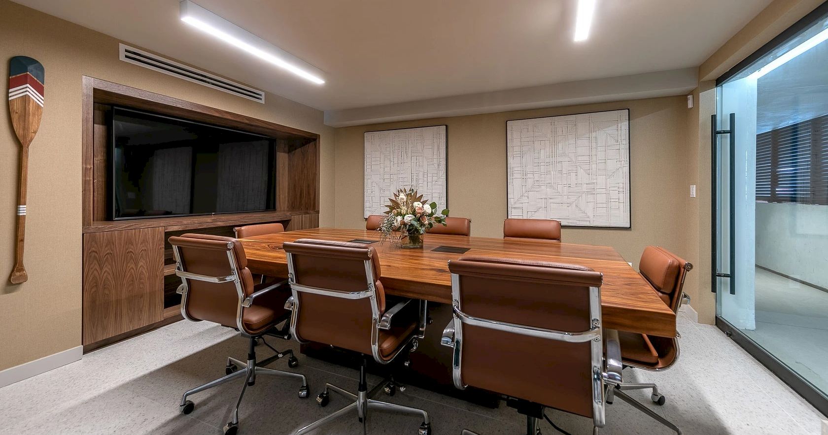 A modern conference room with a long wooden table, brown leather chairs, a TV screen, decor plants, and glass wall to the right.