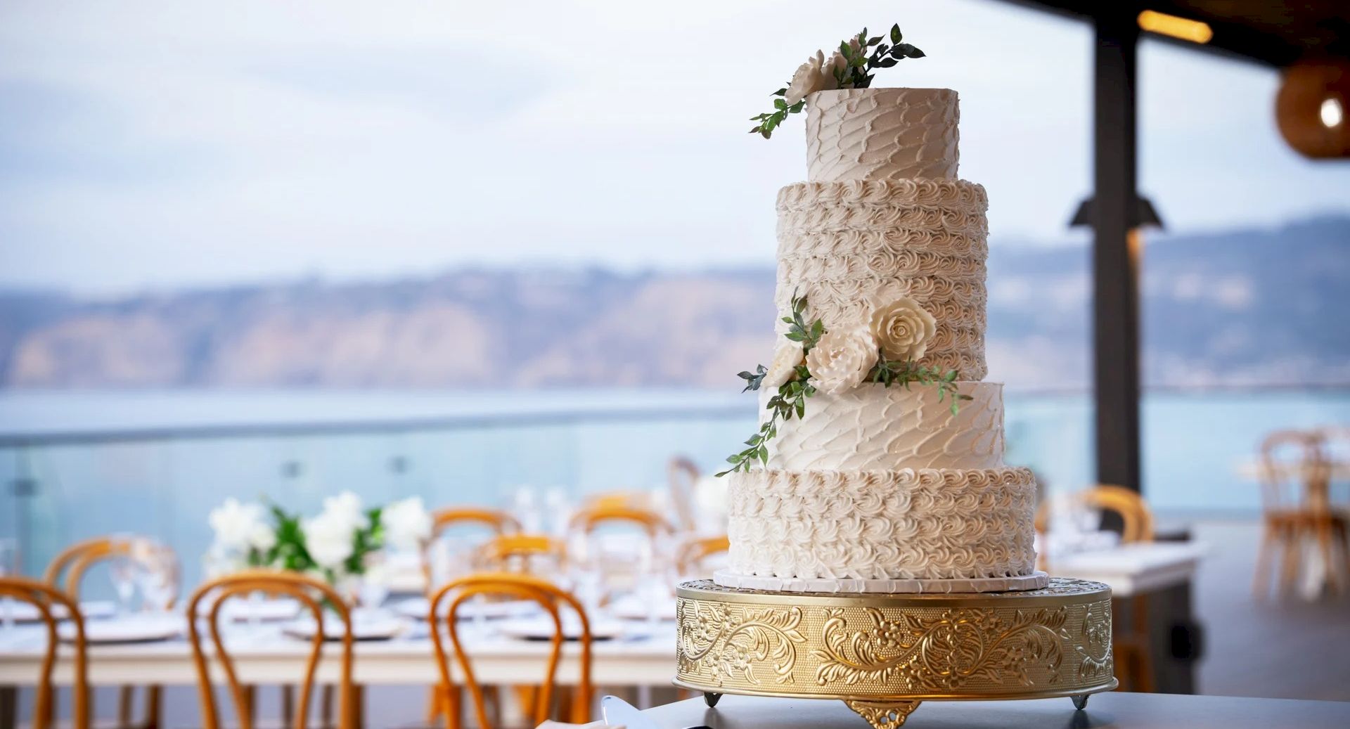 A tall, elegant wedding cake sits on a gold pedestal, adorned with floral accents, at an outdoor venue with ocean views and pastel decor.