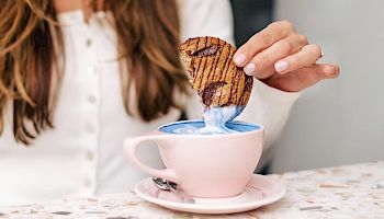 A smiling woman dips a patterned cookie into a blue latte in a pink cup at a cozy table, enjoying a tasty treat.