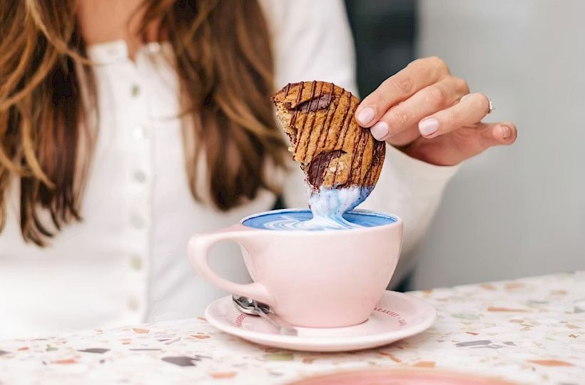A smiling woman dips a patterned cookie into a blue latte in a pink cup at a cozy table, enjoying a tasty treat.