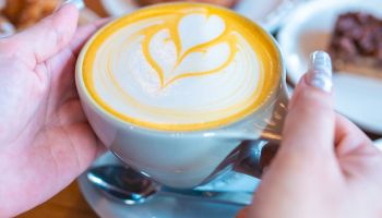 A person holding a cup of latte with a heart-shaped latte art design on top, with pastries and a saucer in the background.
