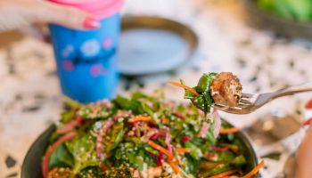 A vibrant salad bowl with greens, sesame seeds, and colorful toppings, served with a pink smoothie in a blue cup and a sunny dining setup.