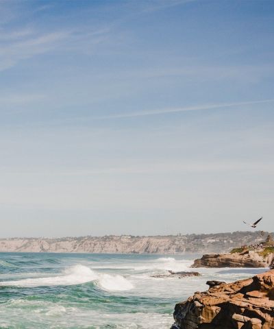 A rocky coastline with waves crashing on the shore, blue sky above, and a bird soaring over the cliffside, ending with a period.
