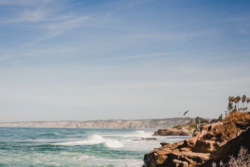 A rocky coastline with waves crashing on the shore, blue sky above, and a bird soaring over the cliffside, ending with a period.