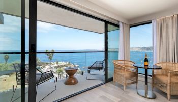 A bright hotel balcony with glass doors opening to a sea view, wicker chairs, a small table, and a bottle of wine nearby, inviting relaxation.