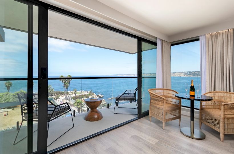 A bright hotel balcony with glass doors opening to a sea view, wicker chairs, a small table, and a bottle of wine nearby, inviting relaxation.