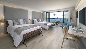 A bright, airy hotel room with two queen beds, soft gray headboards, a seating area, and a balcony overlooking the ocean. Ends cleanly.