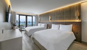 A modern hotel room with two large beds, a sleek desk, and a balcony view of the sea; warm lighting, neutral tones, and clean decor.