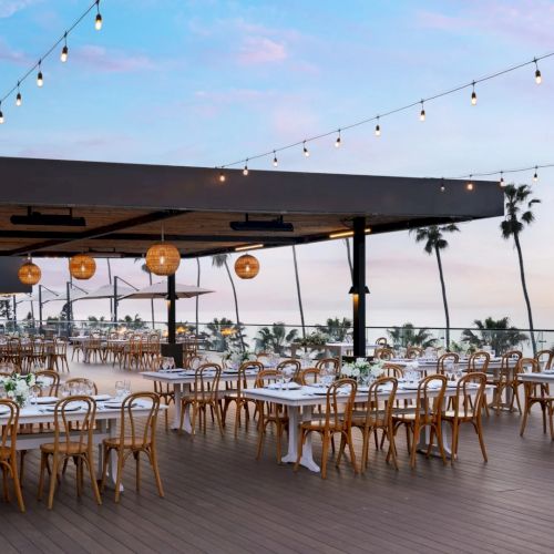 An elegant outdoor beachside dining setup with long wooden tables, string lights, and a shaded pavilion, ready for a seaside feast.