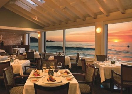 Elegant seaside restaurant interior with white tablecloths, candles, and a sunset view over the ocean. Dining al fresco as waves glow.
