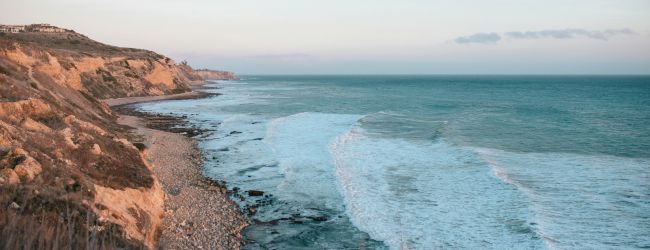 A rocky coastline with cliffs meeting the sea, waves crashing onto a pebble beach under a clear sky.