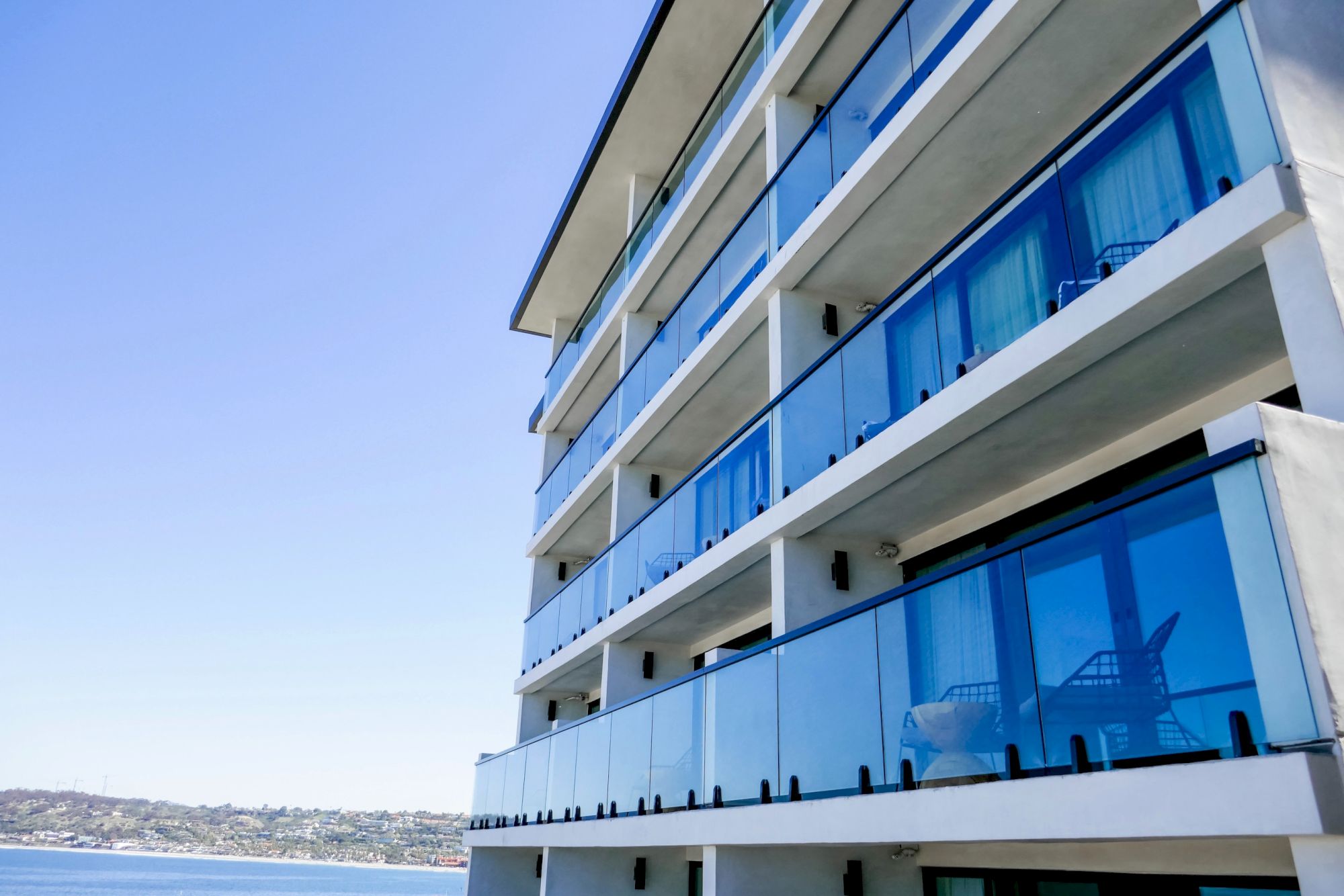 A modern multi-story building with glass balconies overlooking water, bright blue sky in the background.