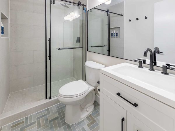 A modern bathroom with a glass-enclosed shower, white vanity, and light gray tile floor, featuring black fixtures and a large mirror.