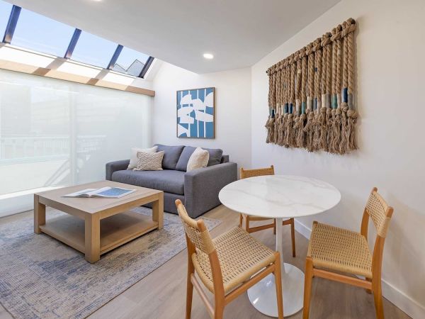 Cozy living area with a gray sofa, coffee table, and a round dining table with woven chairs; sunlight from skylights and a macrame wall hanging.