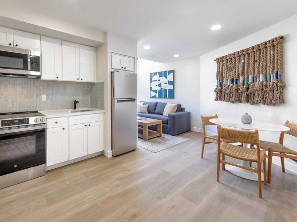 A bright, open kitchen and dining area with white cabinets, stainless appliances, a cozy living nook, and a woven wall hanging.