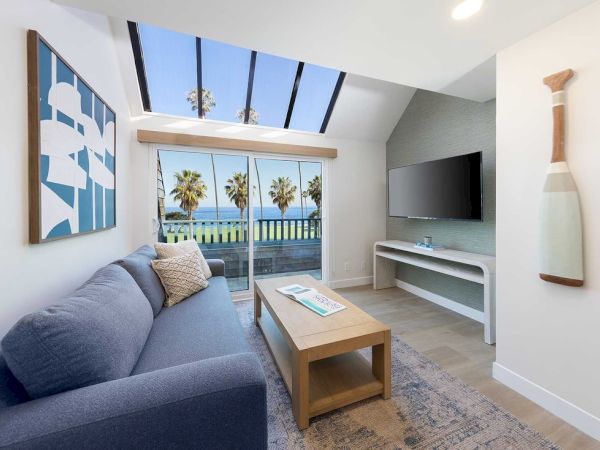Cozy coastal living room with a blue sofa, glass doors to a sunny balcony, a wall-mounted TV, and a beachy paddle decor piece, beach view beyond.