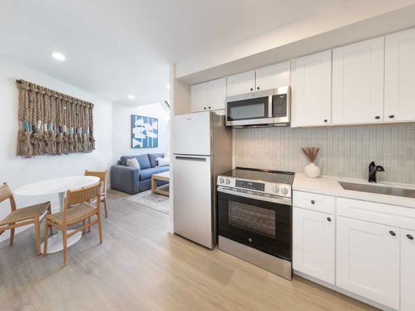 A bright, open kitchen with white cabinets, stainless steel appliances, a small dining table and chairs, and a cozy living area in the background.