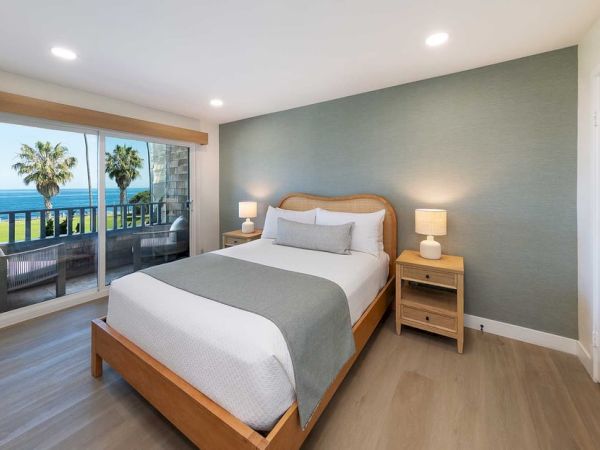 A bright hotel room with a wooden bed, blue accent wall, two nightstands, lamps, and a balcony overlooking palm trees and the ocean.